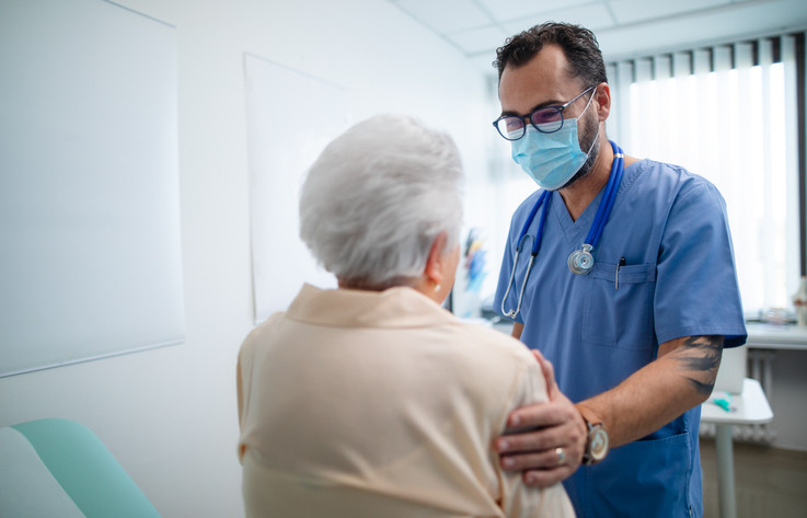 A woman with white hair viewed from behind sitting on an exam table and a doctor wearing blue scrubs and a stethoscope around his neck touching her gently on her upper arm