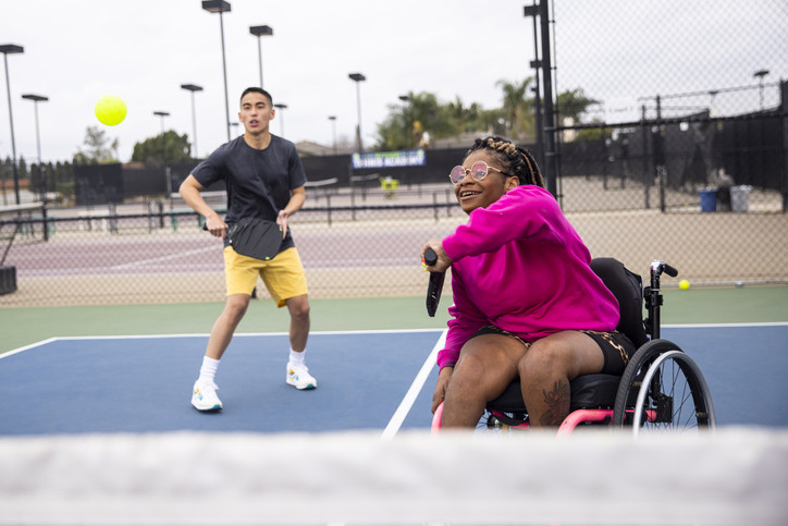 A man on a pickleball court wearing a dark tee shirt and yellow shorts and a woman in a wheelchair swinging back her racquet to hit the ball; she's wearing a bright pink top 
