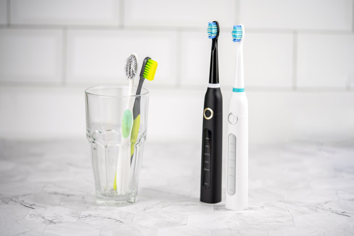 Two electric toothbrushes standing on a marble bathroom counter and two manual toothbrushes in a glass, background is white tile