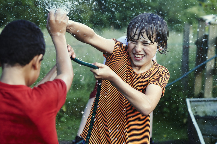 Two boys playing, spraying water on each other with a hose; one wears a red shirt, the other a striped shirt