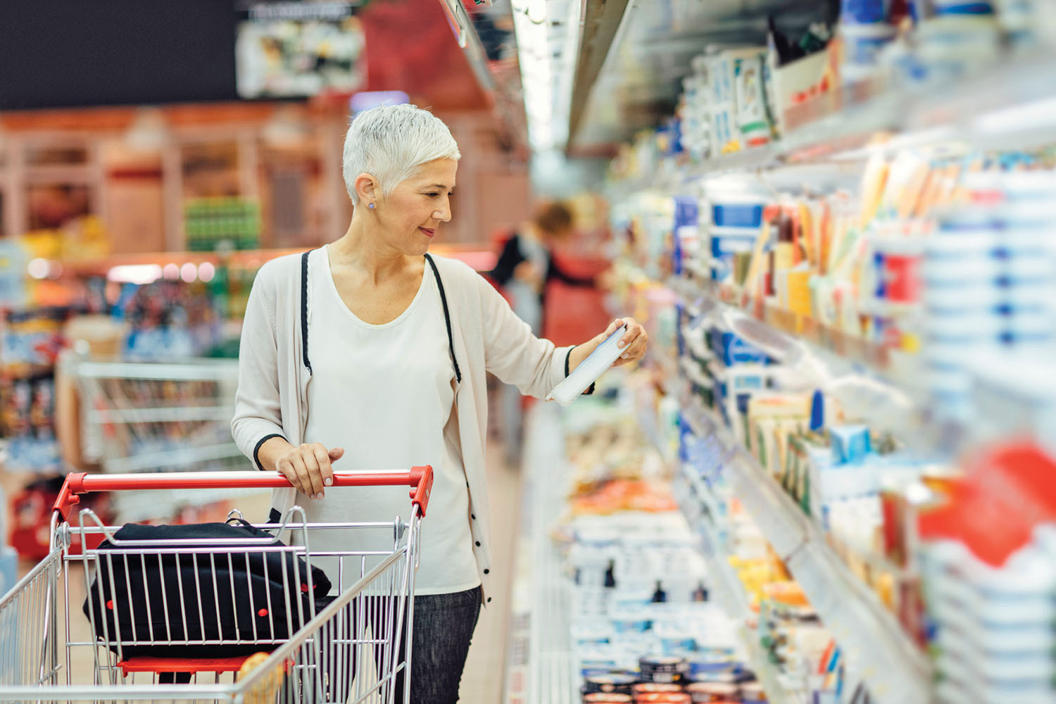 photo of a woman in a supermarket standing behind her shopping cart looking at a food label in the refrigerated section
