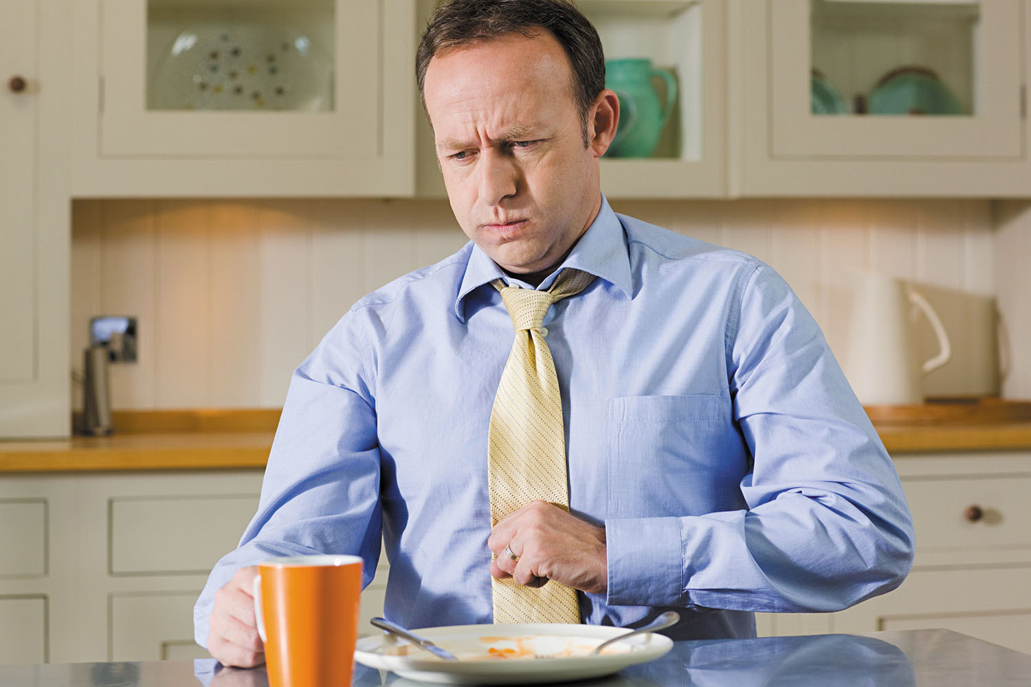 photo of a man who has just finished eating (there is an empty plate in front of him) holding his hand in front of his stomach with a clenched fist and with an expression of distress on his face