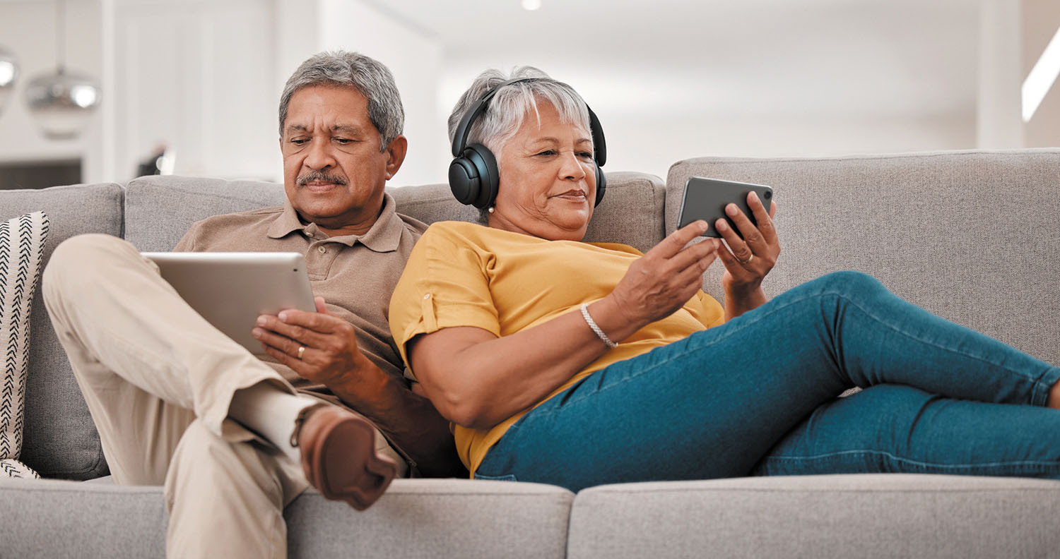 photo of a mature couple sitting on their couch; man is using a tablet while his wife, who is leaning against his shoulder, is looking at her phone and wearing headphones