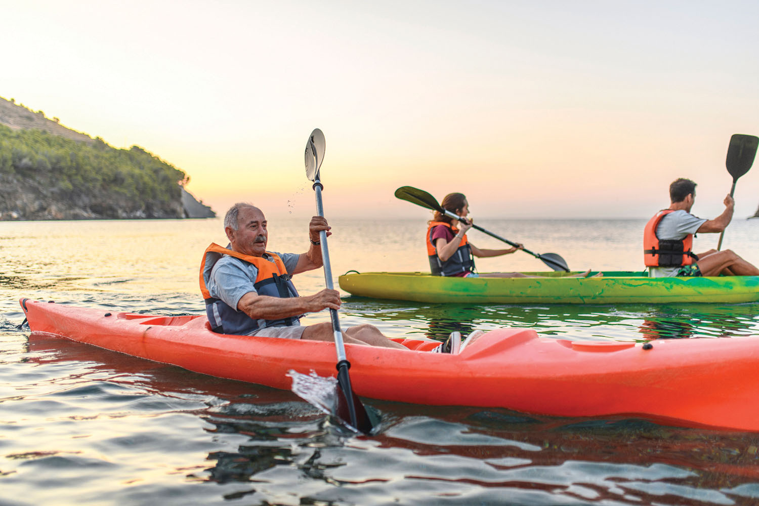 photo of people kayaking, in the foreground is a man by himself in a red kayak and behind him two people in a green kayak