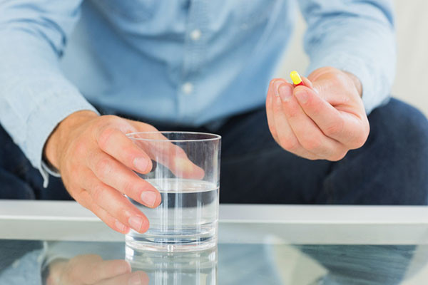 cropped photo shows the torso and arms of a man holding a yellow and red capsule pill in his left hand, right hand is resting on a glass of water