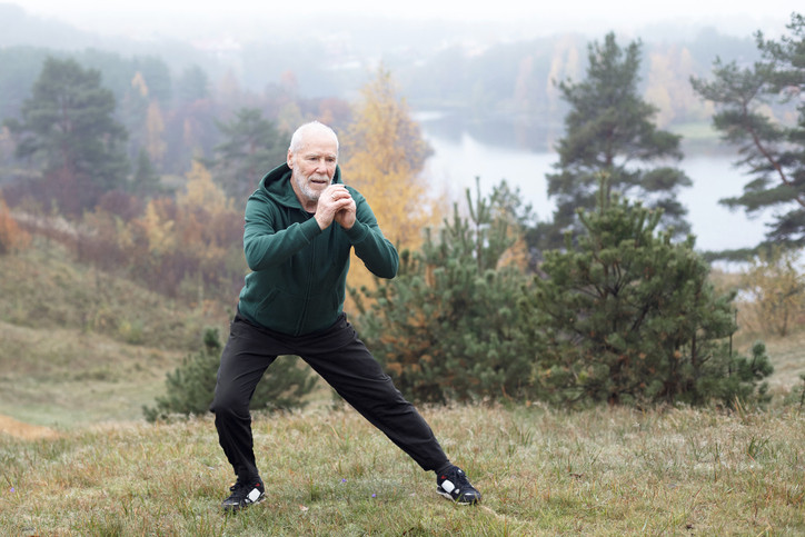 photo of an elderly bearded man in athletic wear warming up before a morning run in a park, misty air and water in the background