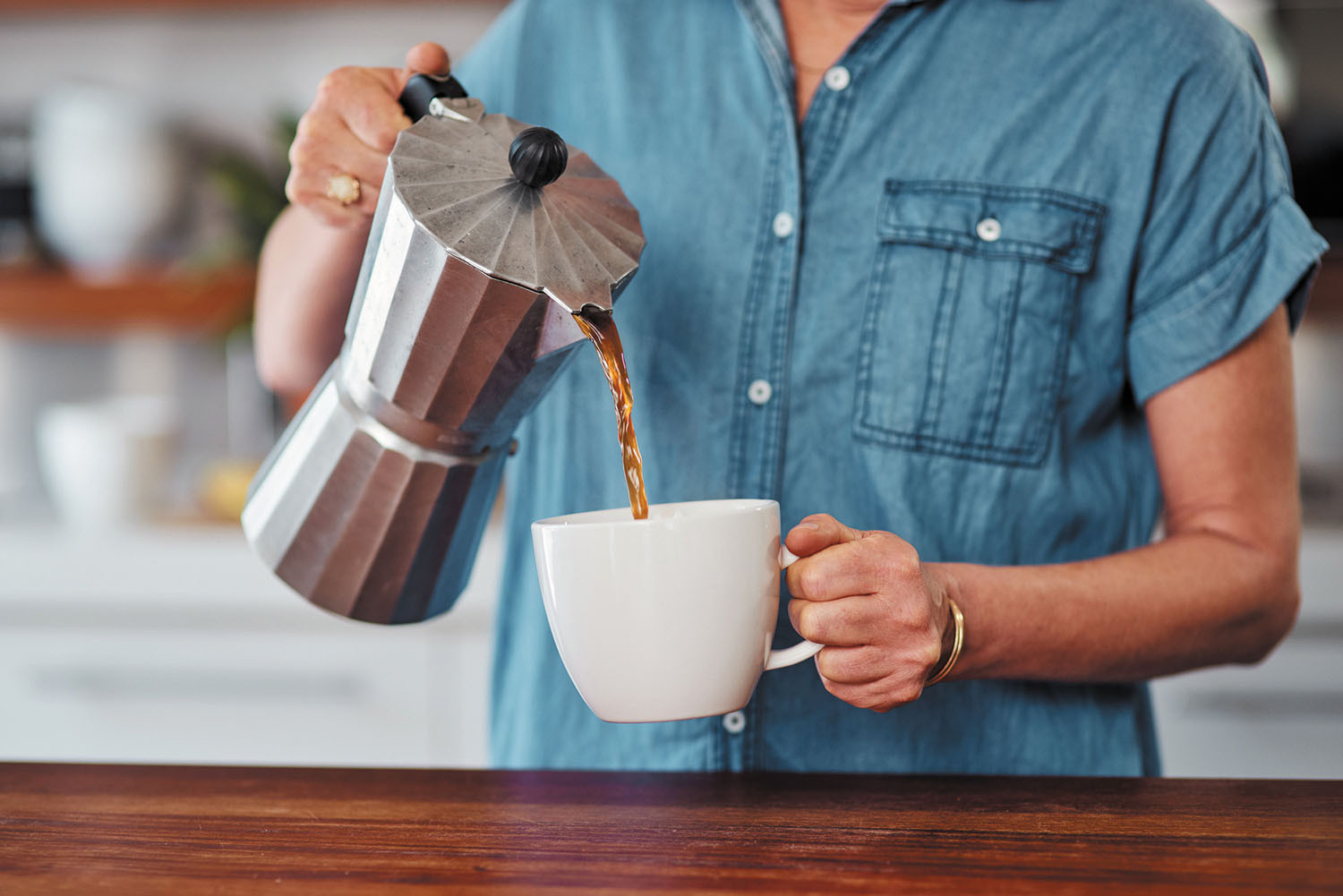 cropped photo showing the torso and arms of a woman pouring coffee from a pot into a white mug