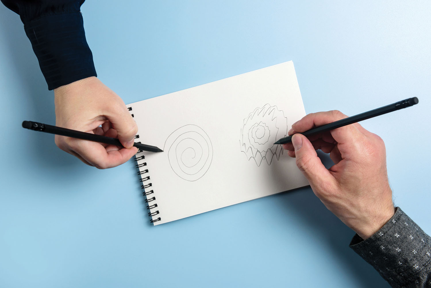 photo of two hands holding pencils and drawing on the same piece of paper, hand on the left has drawn a smooth concentric line while the hand on the right has drawn shakily