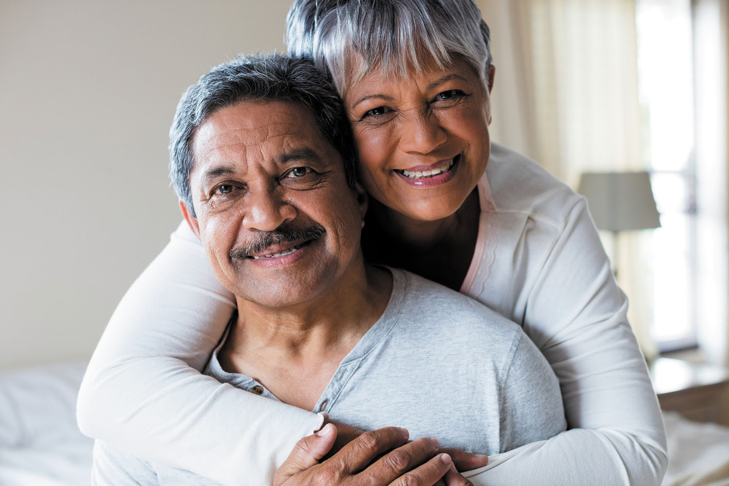 photo of a smiling mature couple, woman is standing behind sitting man and has her arms around his shoulders and chest