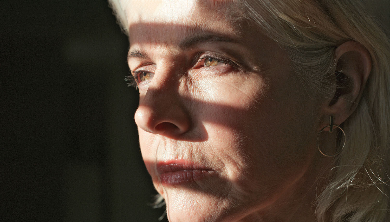 close-up photo of the face of a somber woman, light and shadow from a nearby window are shown across her face