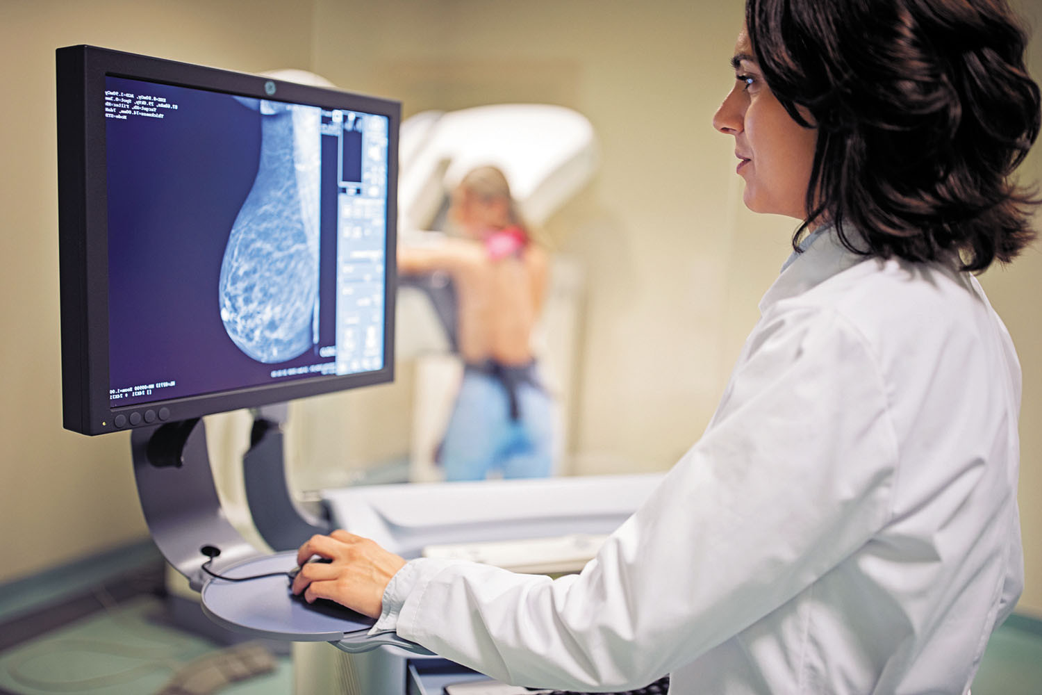 photo of a medical technician viewing mammogram on a computer screen; in the background, out of focus, can be seen the woman being examined with her back to the camera