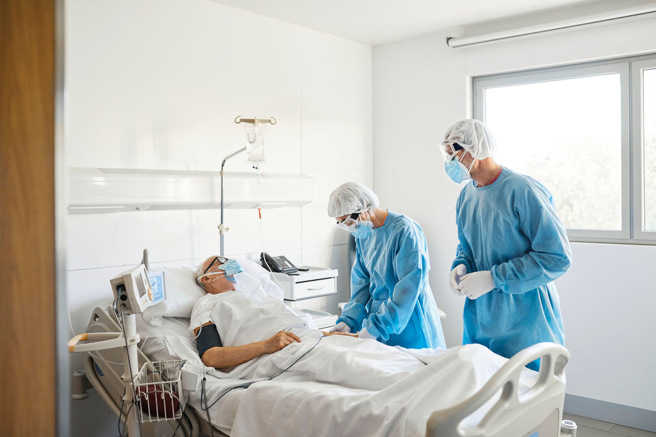 photo of a mature patient in a hospital bed being tended to by two providers wearing protective gear