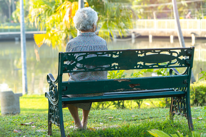 photo of an elderly woman viewed from behind, sitting on a bench in a park with fall foliage visible in the background
