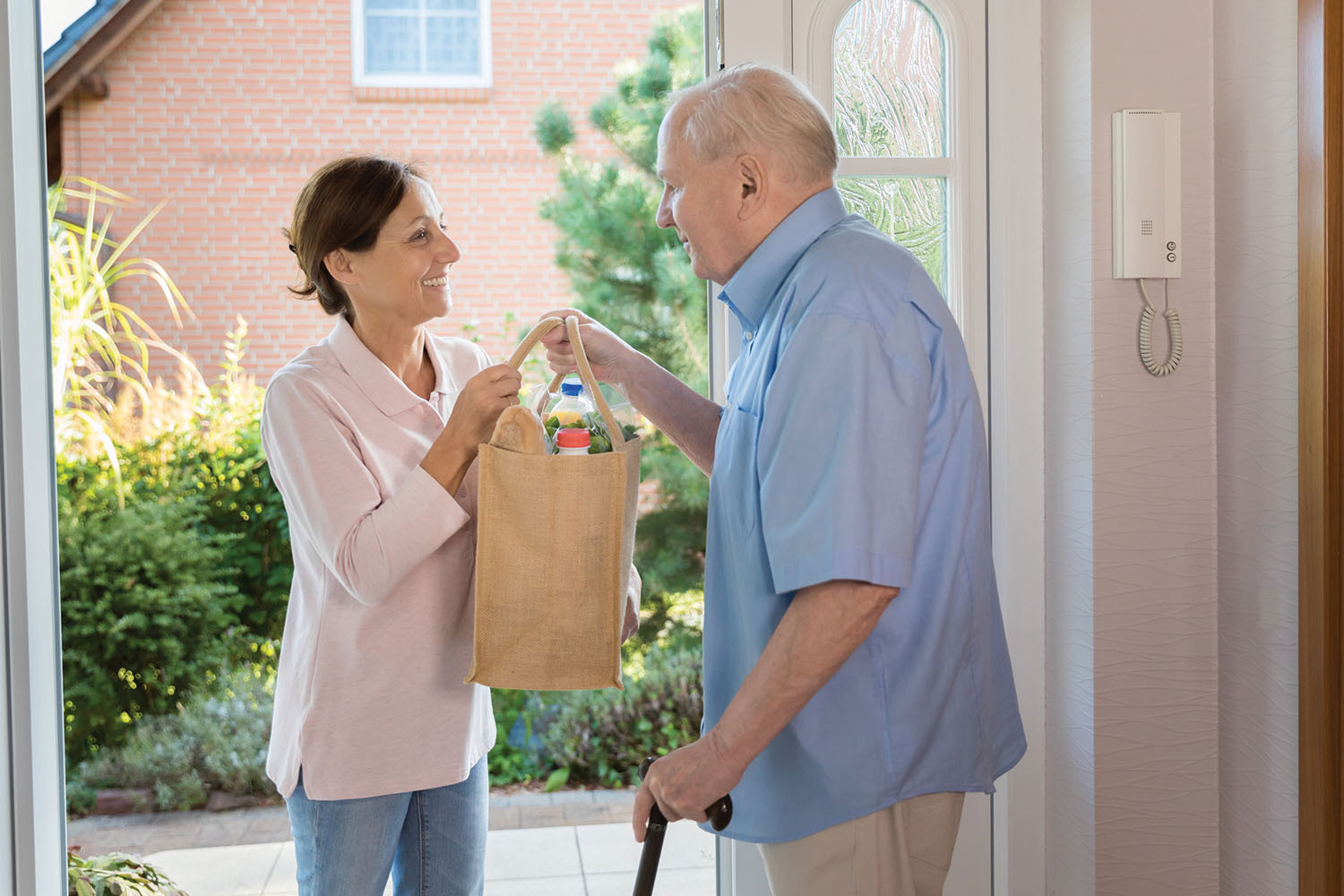 photo of a smiling woman handing a bag of groceries to a man on the front porch of his home, the man has a hearing aid visible in his ear and is using his other hand to lean on a cane