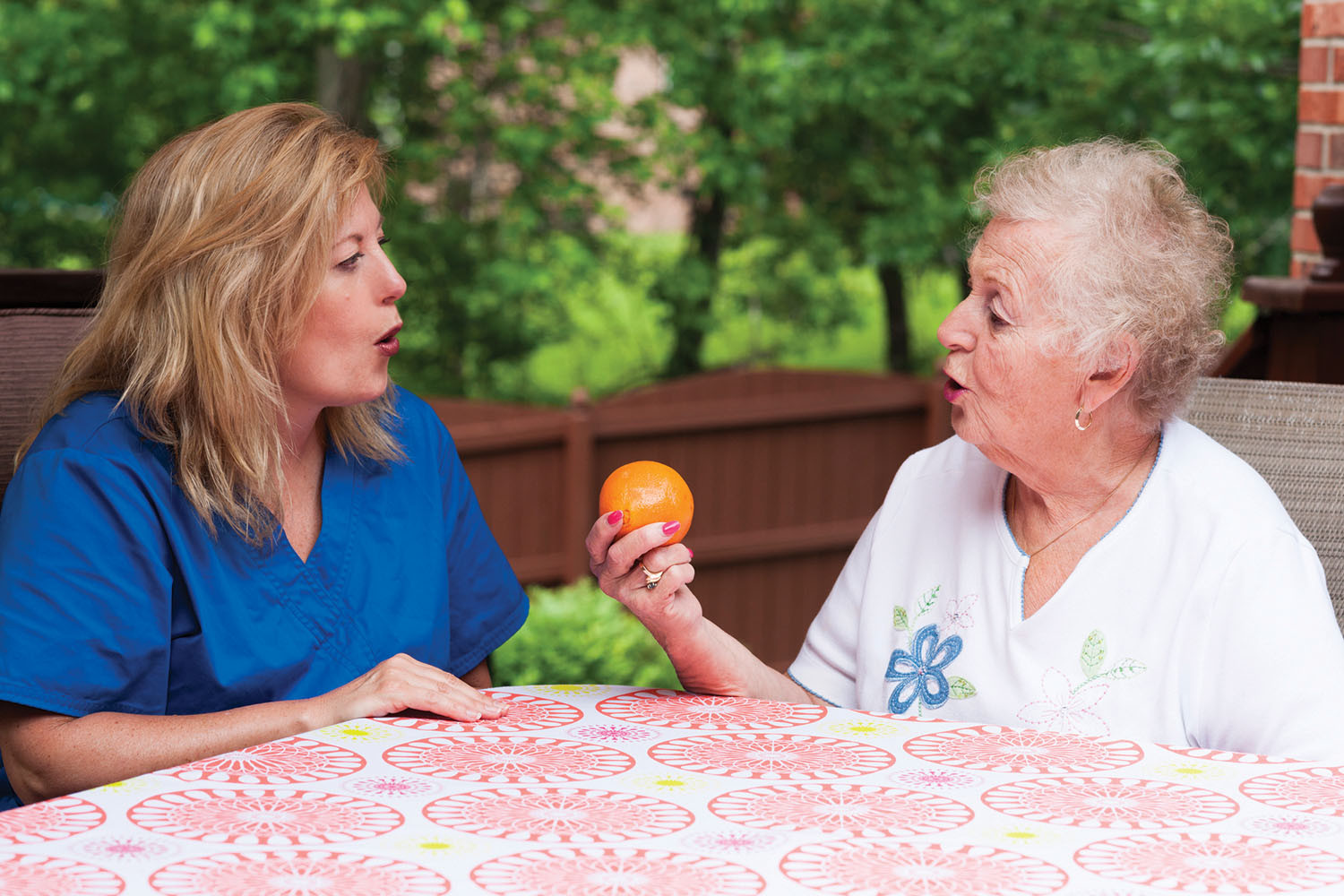 photo of a senior woman holding an orange while working with a speech therapist, they are sitting at a table facing each other