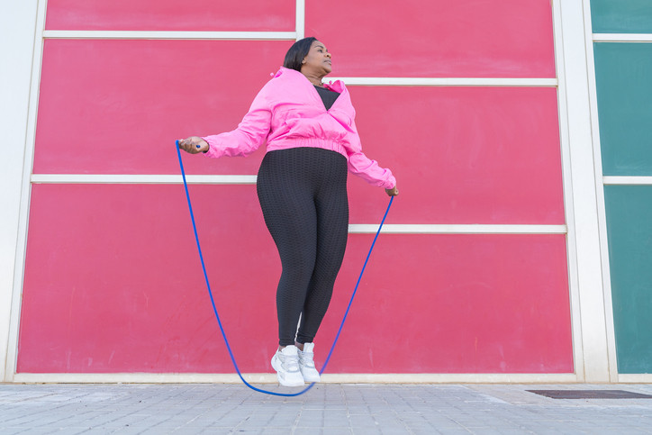 Woman jumps rope a few inches above gray bricks, wearing pink jacket and black leggings, pink rectangle background; concept plyometrics