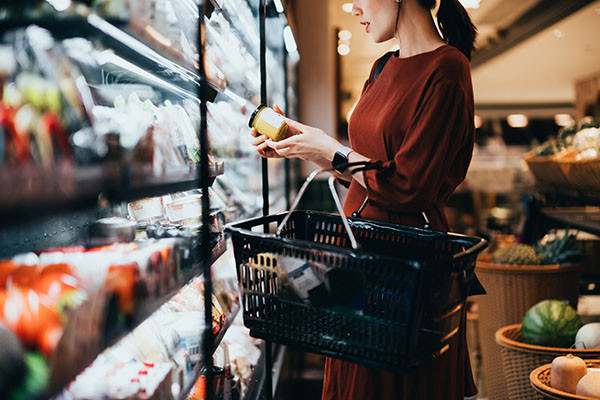 Photo of a young woman with a shopping basket on her arm, standing in the dairy aisle, reading the label on a jar