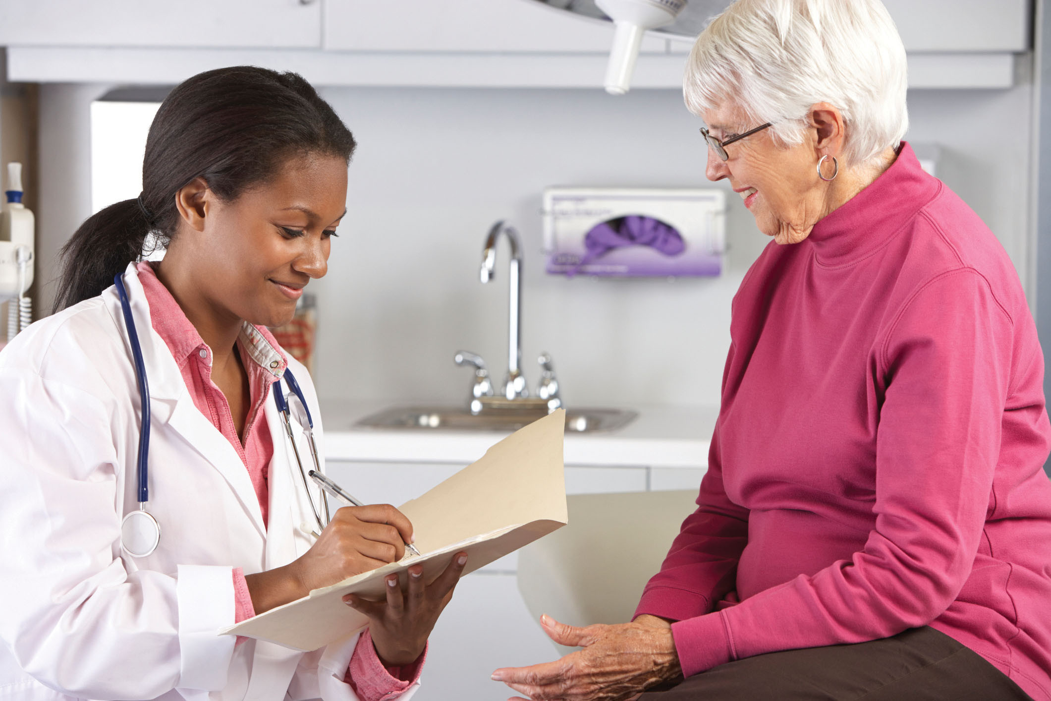 photo of a medical professional talking with a senior woman in an exam room; she is holding a folder and writing on paper in it