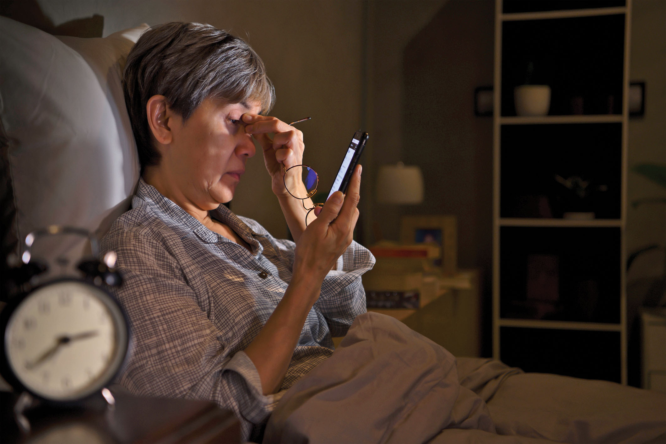 photo of a woman in bed in a dark room, using her smartphone and rubbing her eyes; clock on the night table shows 2:40 AM