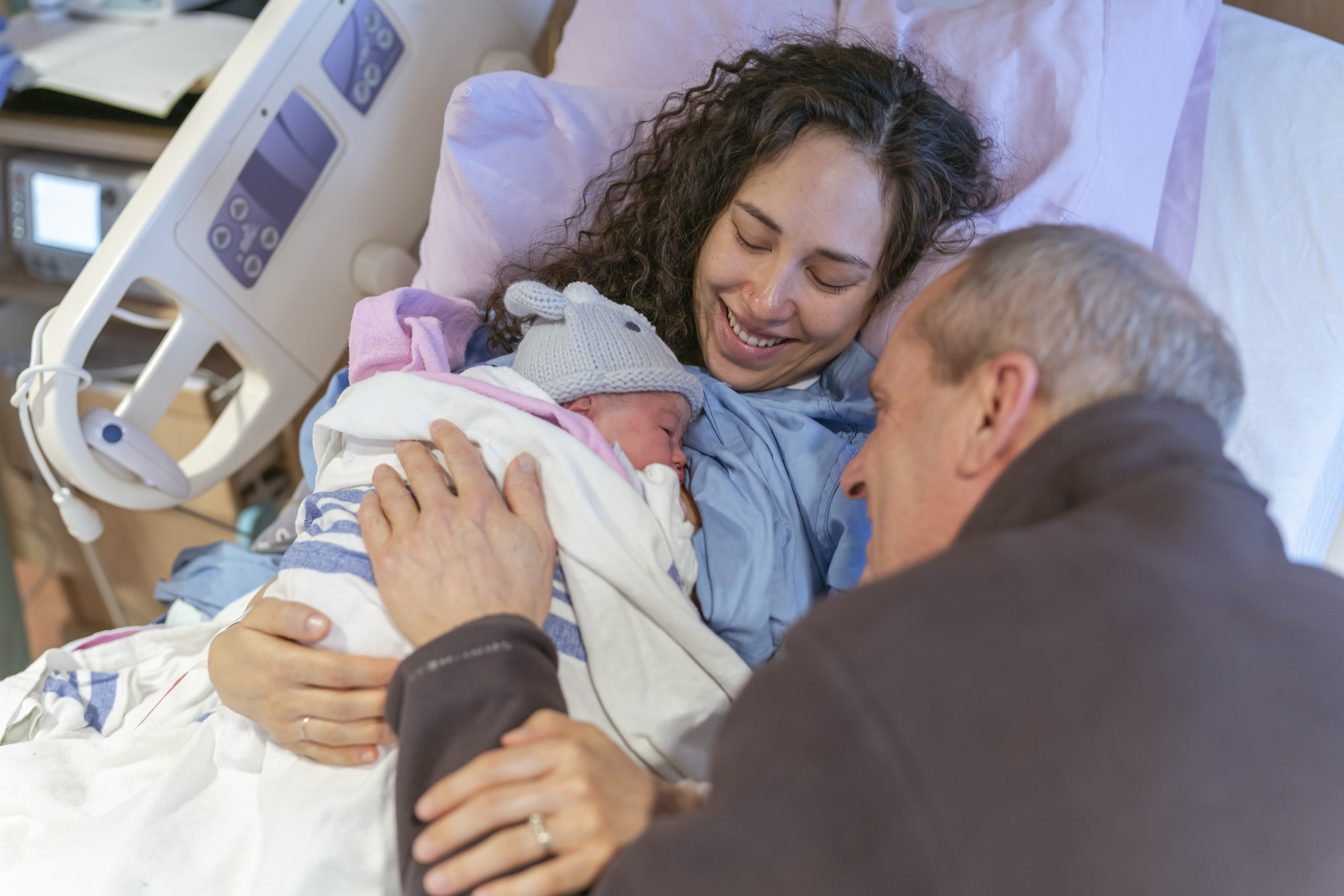 A photo of a caucasian grandpa who is emotional and happy while seeing his newborn granddaughter for the first time in the hospital room. He is looking at the baby and smiling while his daughter and the baby's mother are lying down together.