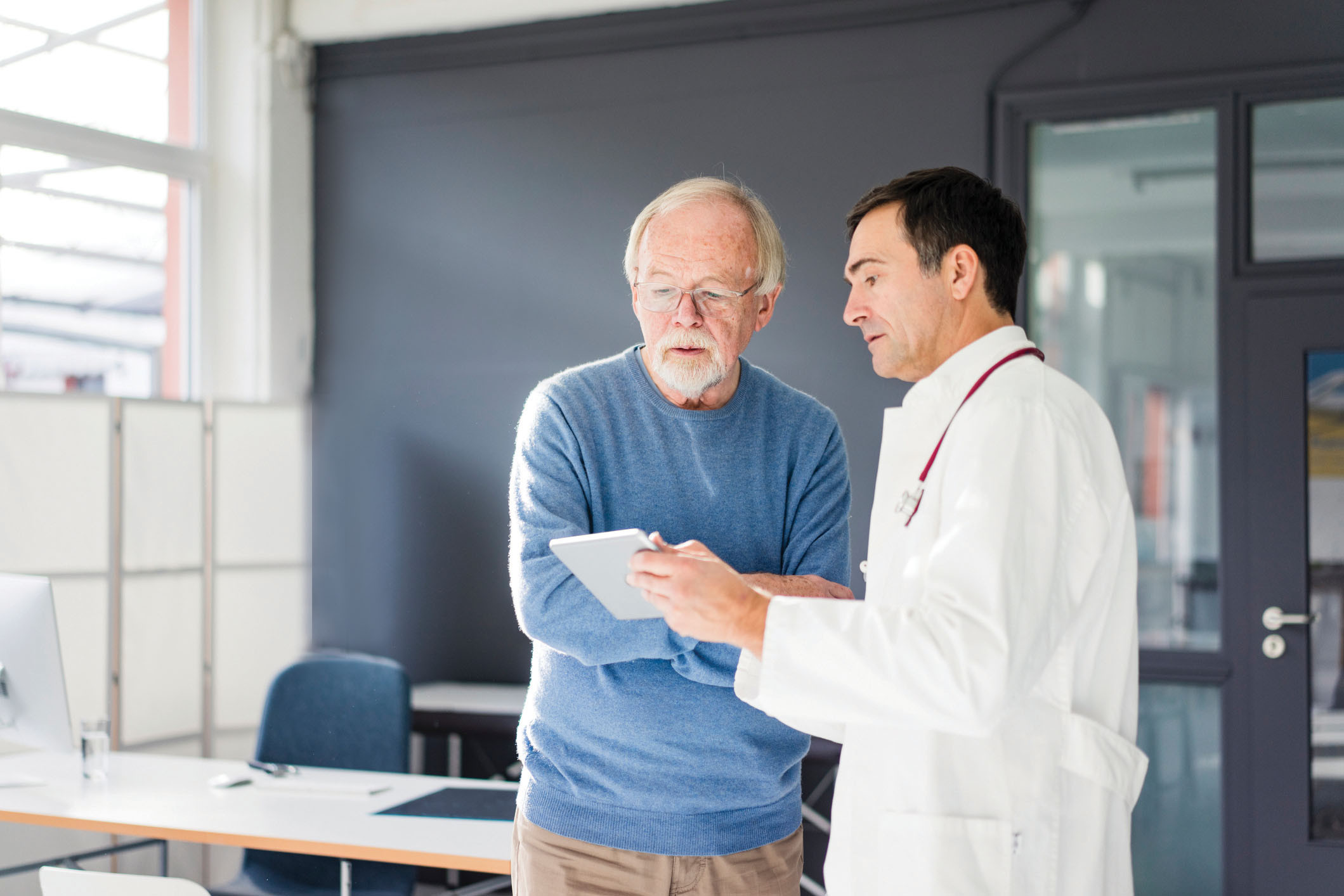 photo of a doctor and patient having a conversation while standing; the doctor is holding a tablet and showing it to the patient