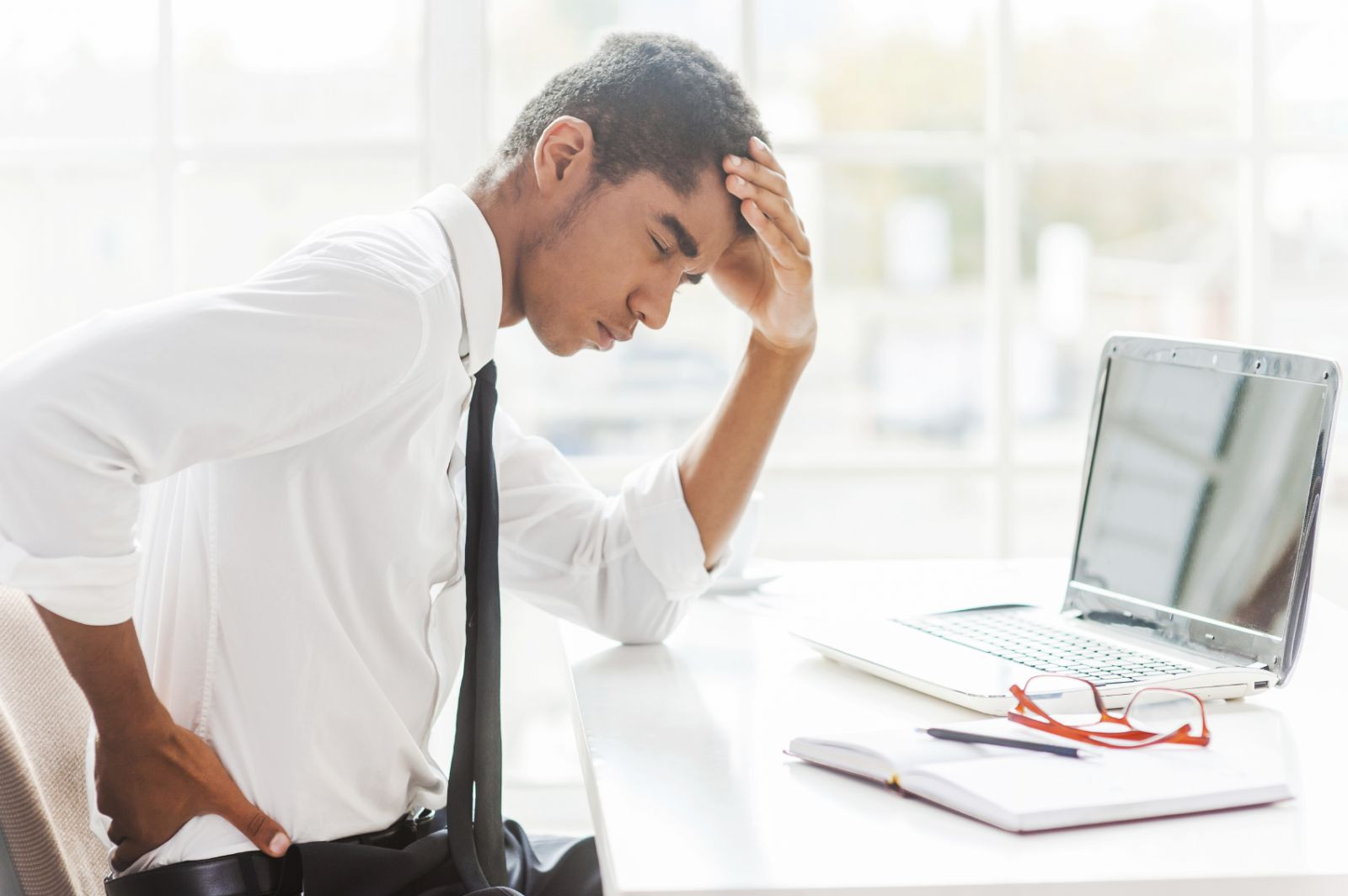 photo of a man sitting at a desk holding one hand on his lower back; the other is resting on his forehead