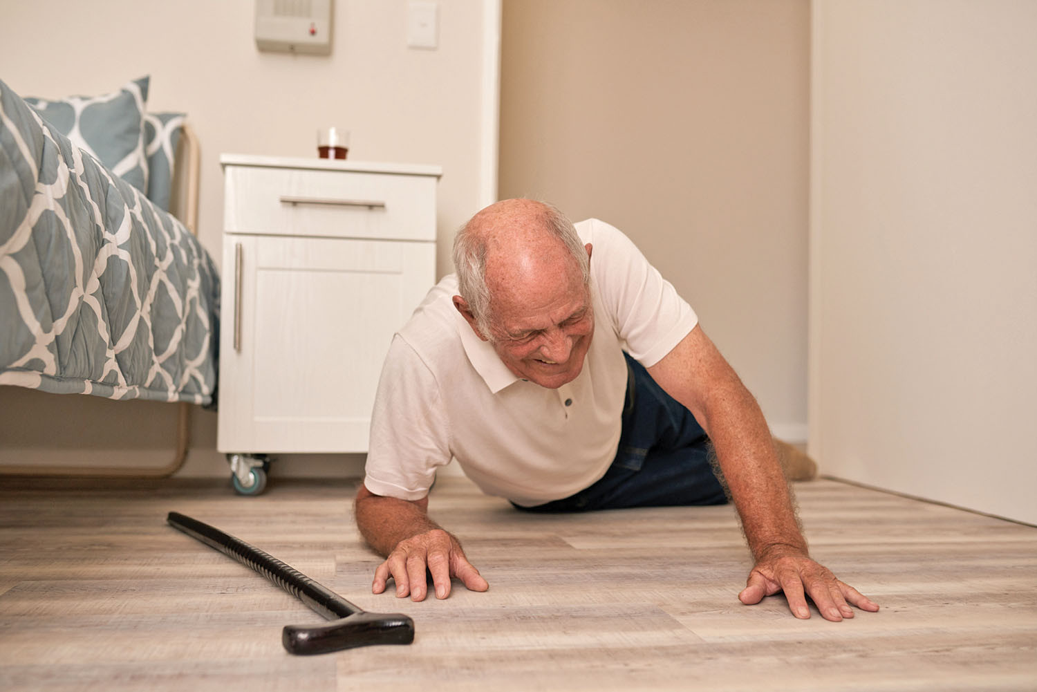 photo of a senior man who has fallen in his bedroom, cane is on the floor next to him and he is trying to push himself up while grimacing in pain