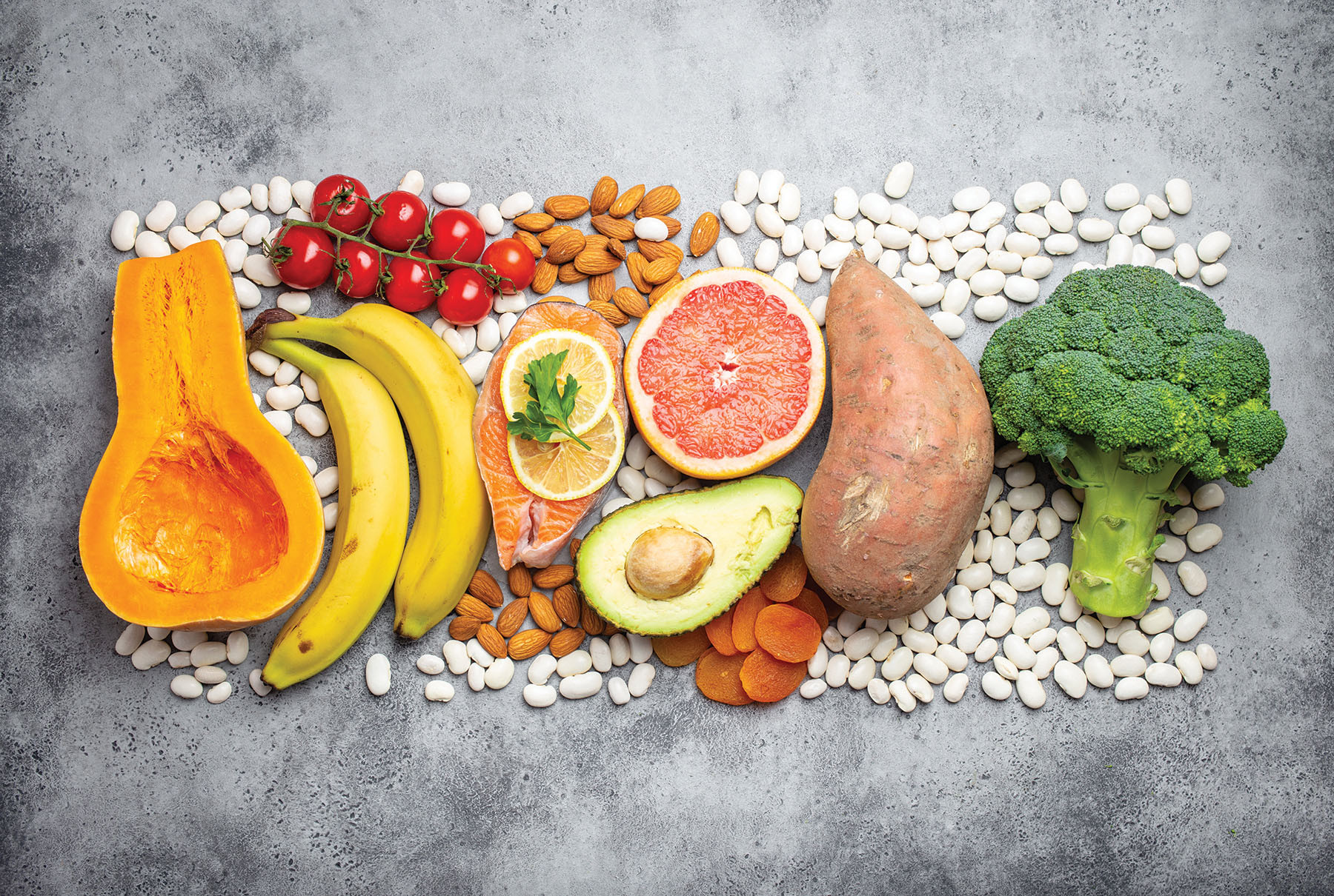 photo of an assortment of foods that are good sources of potassium, including swweet potato, bananas, white beans, grape tomatoes, broccoli, avocado, and salmon