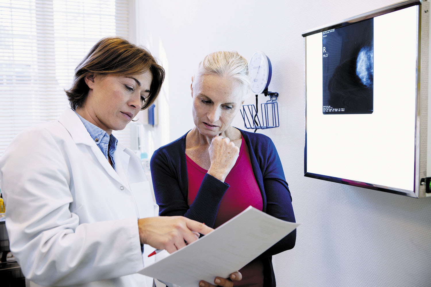 photo of a doctor showing patient the results of a test; doctor is holding papers in her hand, and there is a light box on the wall next to them showing a mammogram