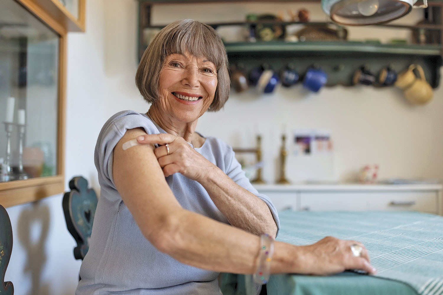 photo of a smiling woman sitting in her kitchen, pointing to a bandage on her upper arm where she received a vaccination