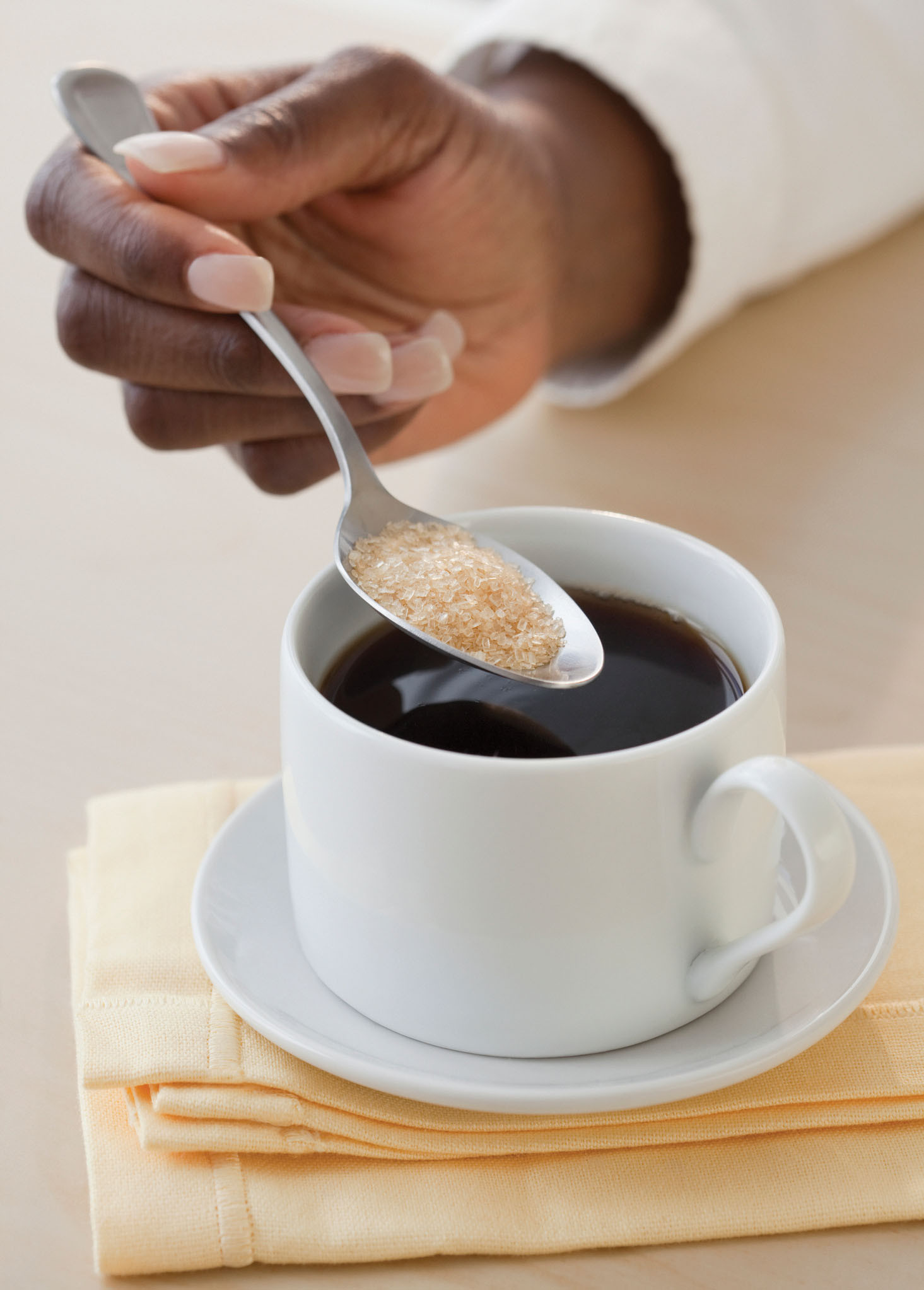 photo of a hand holding a spoon of raw sugar ove a cup of black coffee
