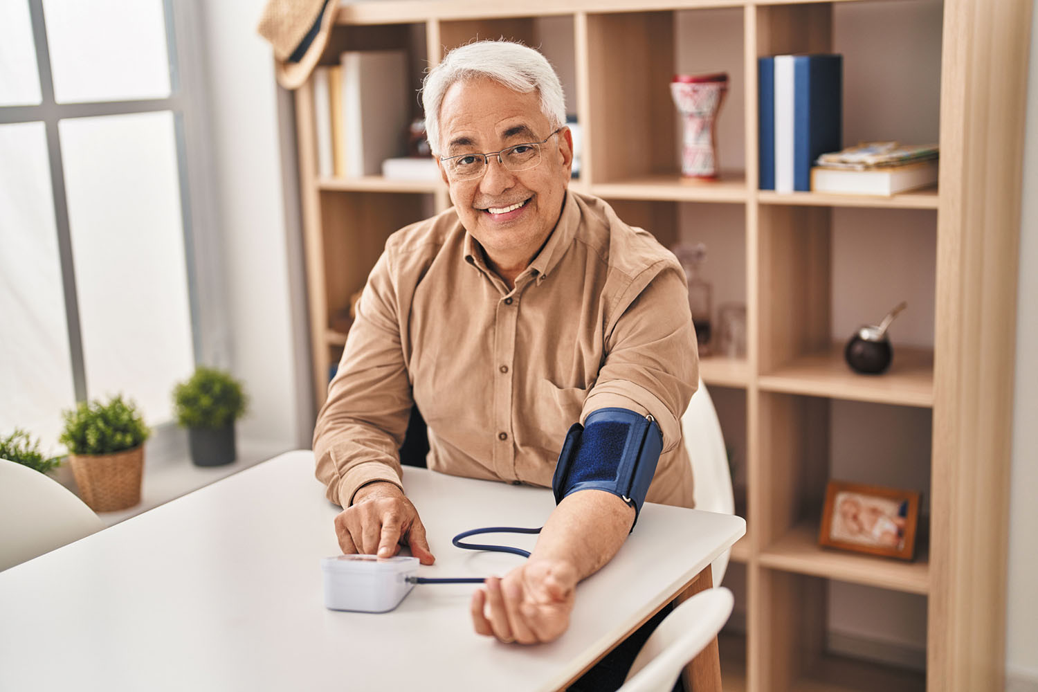 photo of a senior man seated at a table and smiling as he takes his blood pressure using a home monitor