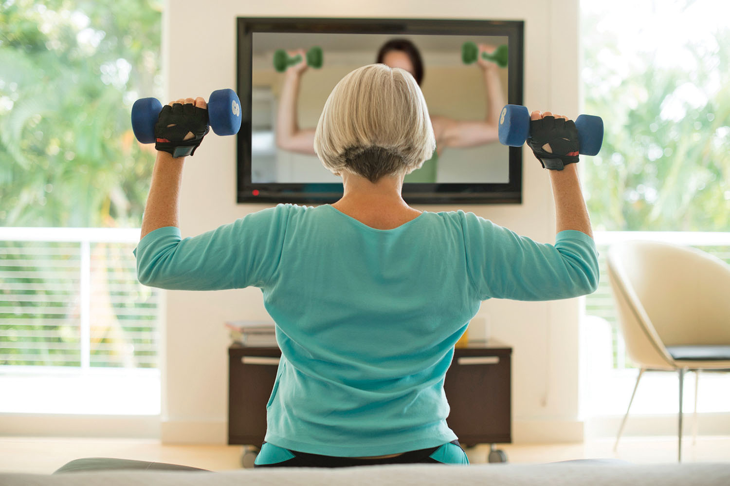 photo of a woman viewed from behind as she exercises with hand weights in her home while watching a virtual exercise class on her TV