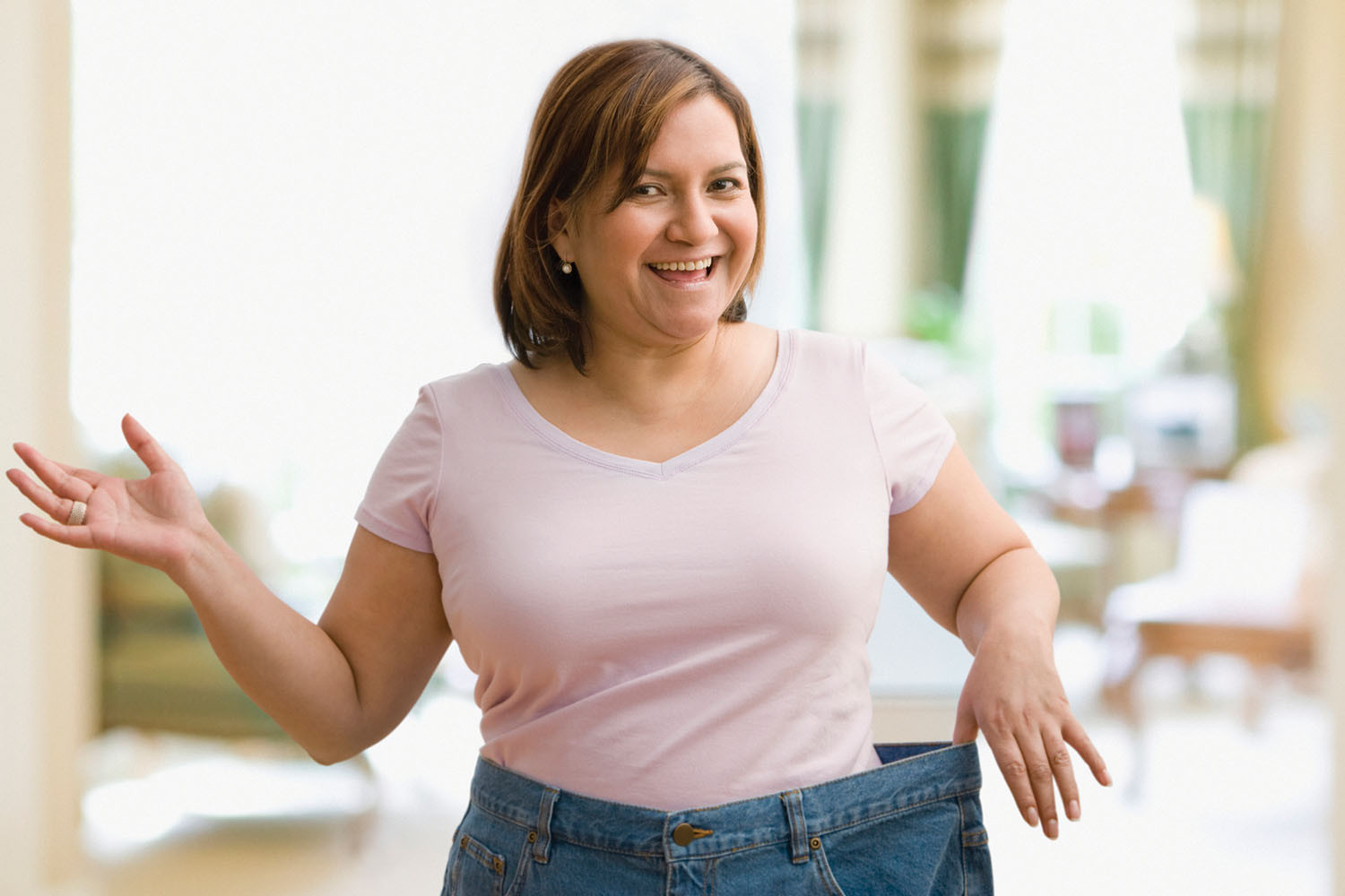 photo of a smiling woman showing off weight loss by holding out the waist of the pants she is wearing that are now too big for her