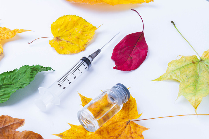 Yellow, red, and greenish autumn leaves with a vaccine syringe and vial posed against a white background; concept is fall vaccines