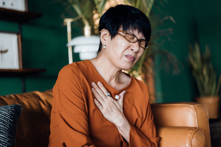 A photo of a senior Asian woman with eyes closed holding her chest in discomfort, suffering from chest pain while sitting on sofa at home.
