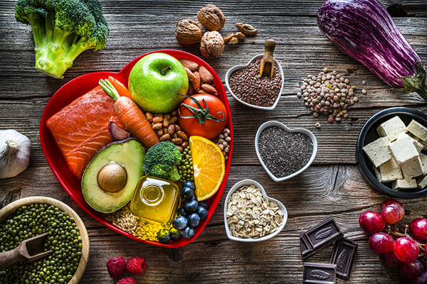 photo of an assortment of heart-healthy foods arranged and displayed on a weathered wood table: salmon, broccoli, apple, dishes of beans and peas, avocado, eggplant, nuts, tofu, oats, dark chocolate, and more