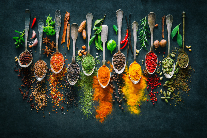 Colorful herbs and spices arrayed in sprays and heaped on silver teaspoons against a dark background