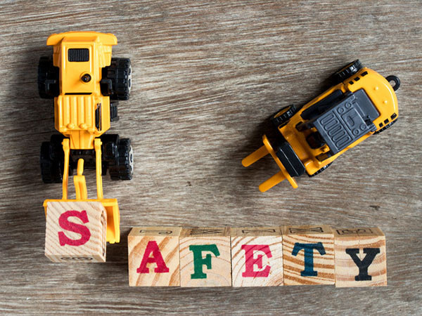 A yellow and black toy bulldozer lifts the letter "s" in wooden blocks spelling out safety; a toy forklift is nearby