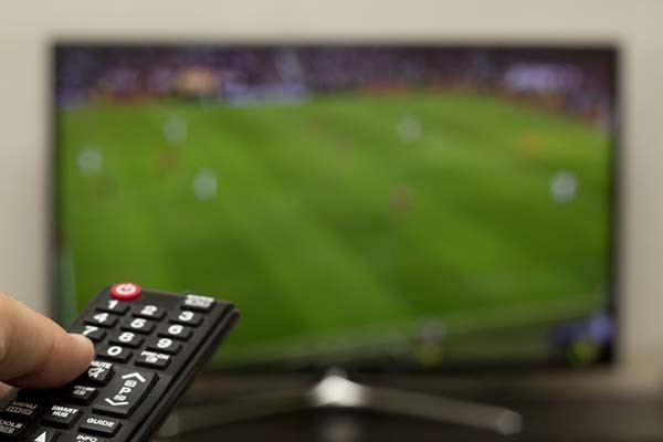 photo of a hand holding a remote control at the left edge of the image; behind it is a TV that is out of focus, but a soccer game can be discerned on the screen