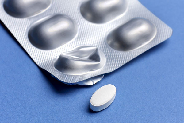 close-up photo of a blister pack of pills on a blue background with one oval pill pushed out of the packaging and positioned in front of the pack