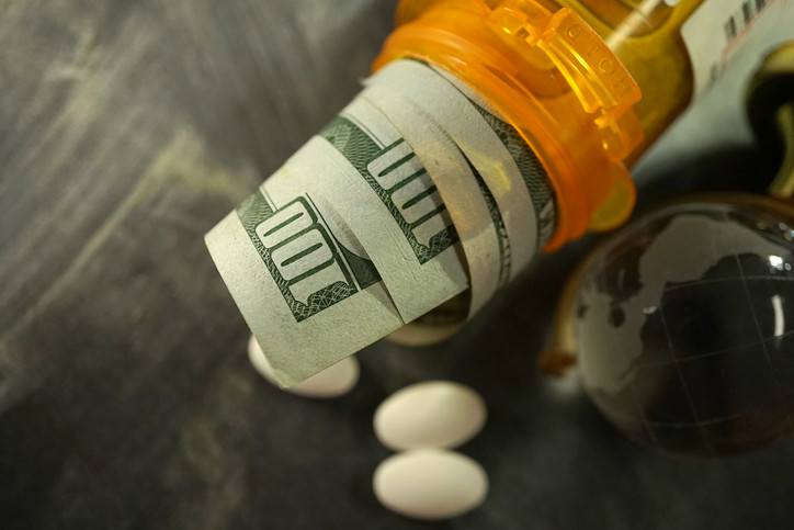 Orange plastic prescription drug bottle tipped on its side with several rolled $100 bills tucked inside peeking out; a few white oval pills & glass marble globe lie next to it