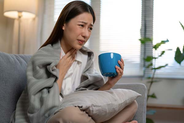 photo of a sick woman sitting on a couch in visible discomfort; she is holding a blue mug with one hand and the other to her throat