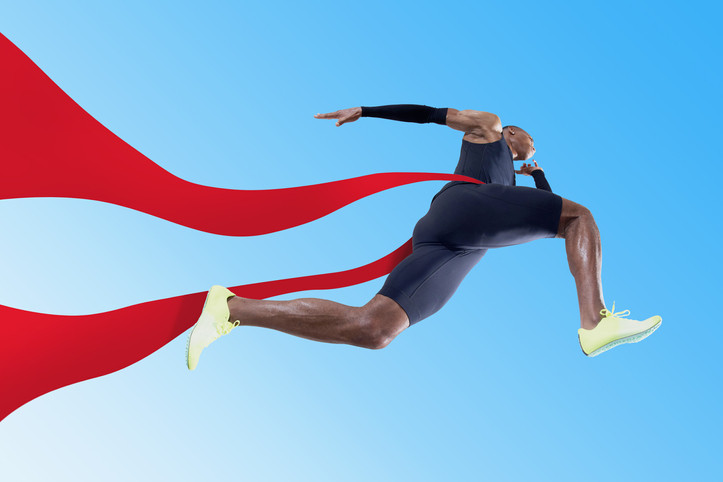 A well-muscled runner in black workout gear and greenish-white sneakers pictured leaping from below crossing a red ribbon finish line against a sky blue background 