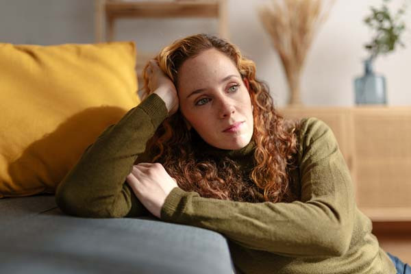 photo of a woman sitting on the floor with her elbow on a sofa cushion and hand on the side of her head, staring dreamily lost in her thoughts