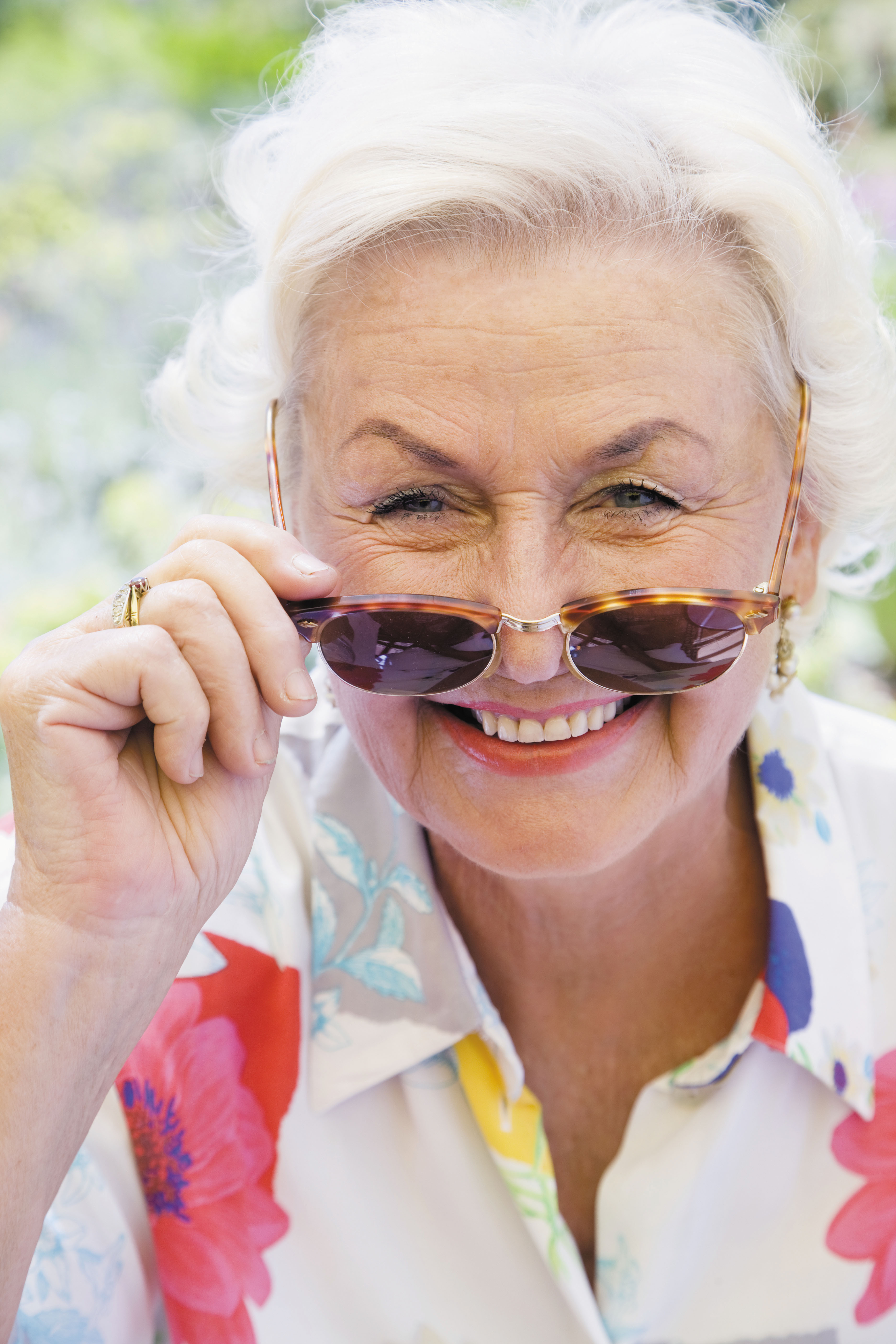 photo of a smiling older woman holding sunglasses that have been pulled down her nose so her eyes are visible