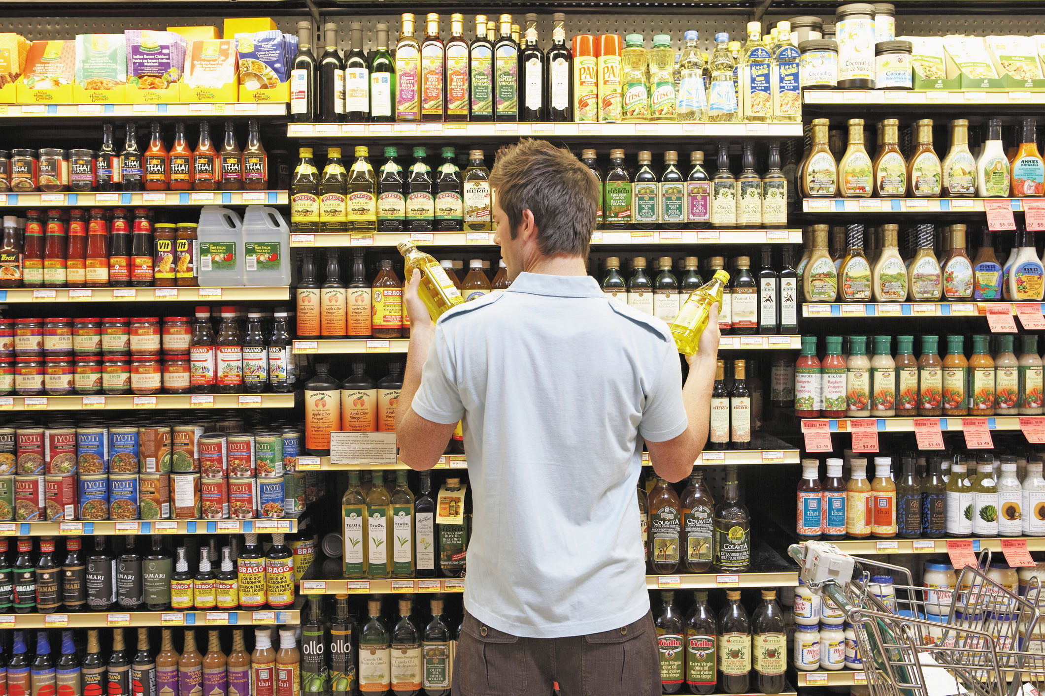 photo of a man viewed from behind as he stands in a supermarket aisle and compares two bottles of cooking oil, one in each hand