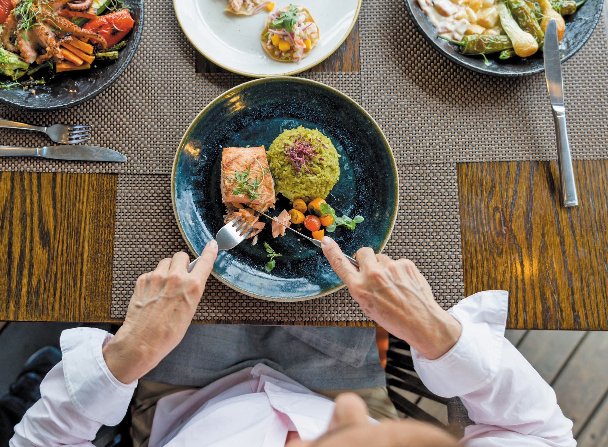 cropped photo overhead view of a person's hands using a fork and knife to cut a piece of salmon; vegetables and couscous are also on the plate
