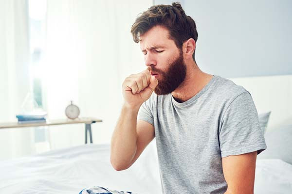 photo of a man sitting on a bed coughing, holding his hand in front of his mouth