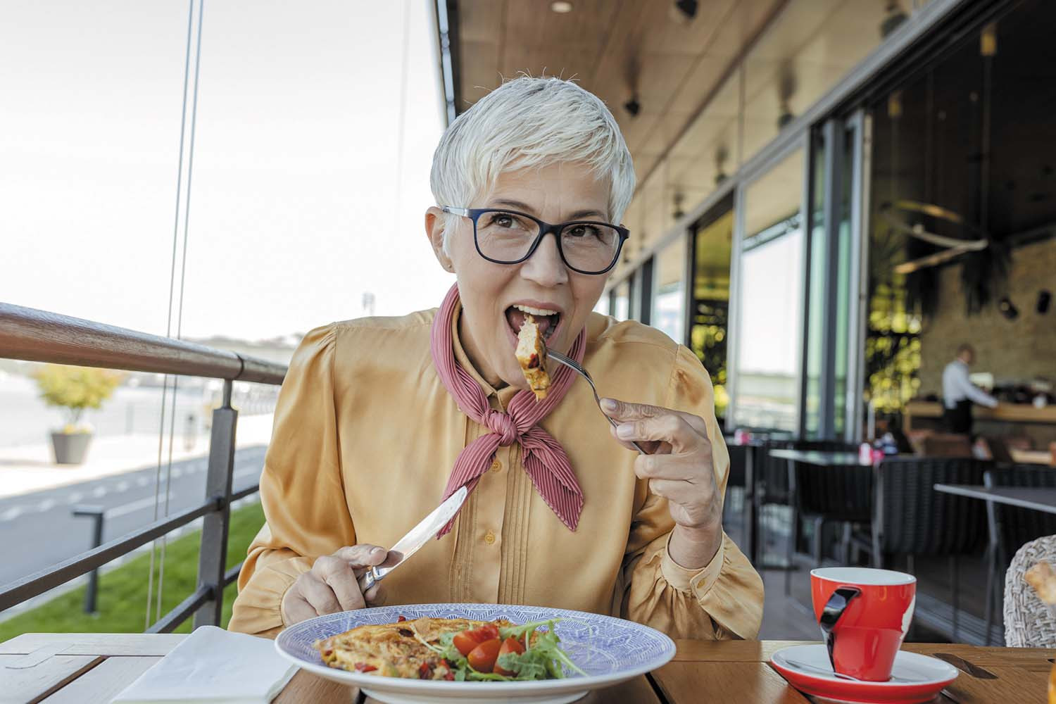 photo of a senior woman at a table on an outdoor balcony, holding a fork and about to put a bite of chicken into her mouth